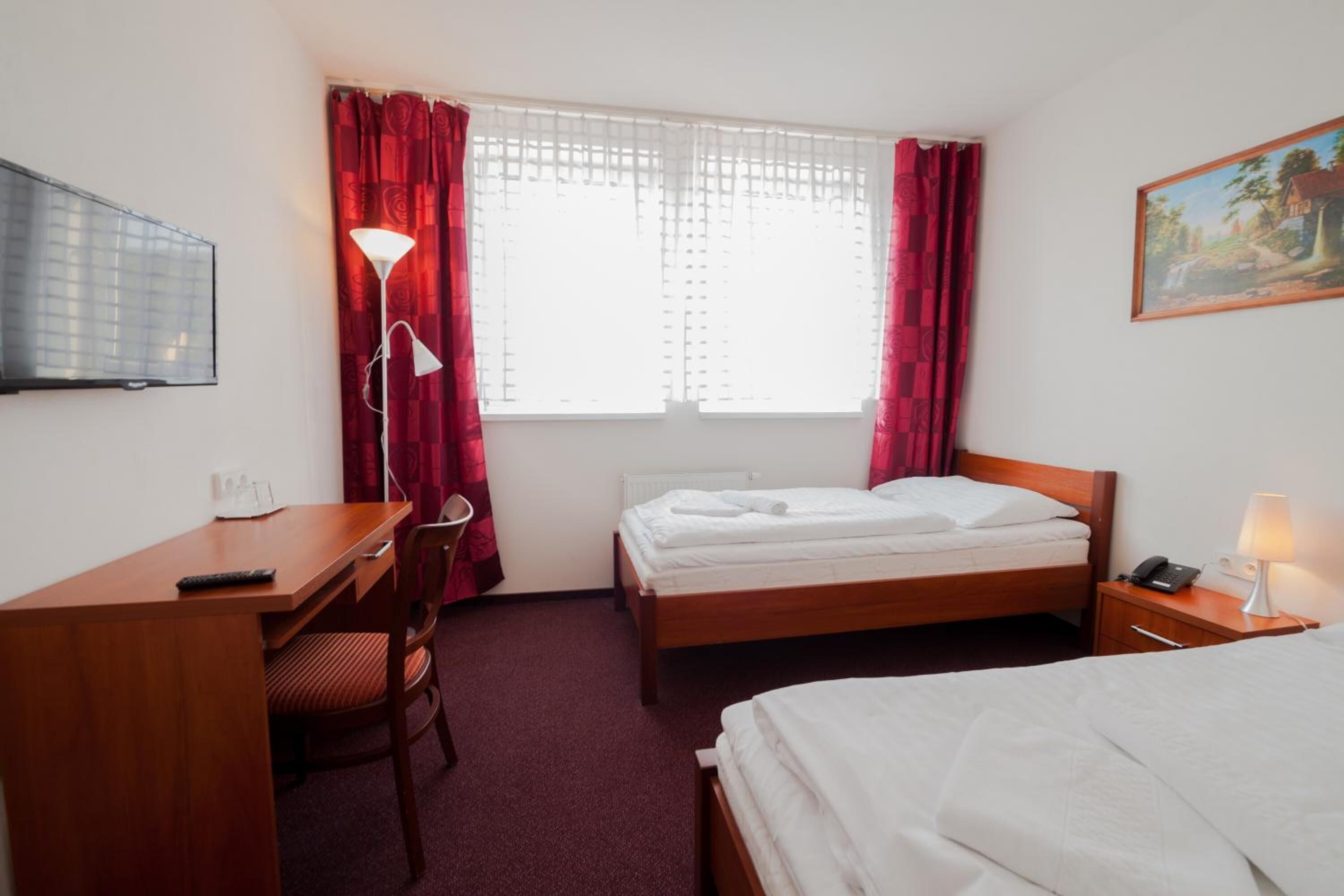 A hotel room with a single bed, a desk with a chair, a bedside table with a lamp, a TV on the wall, and a window with curtains.