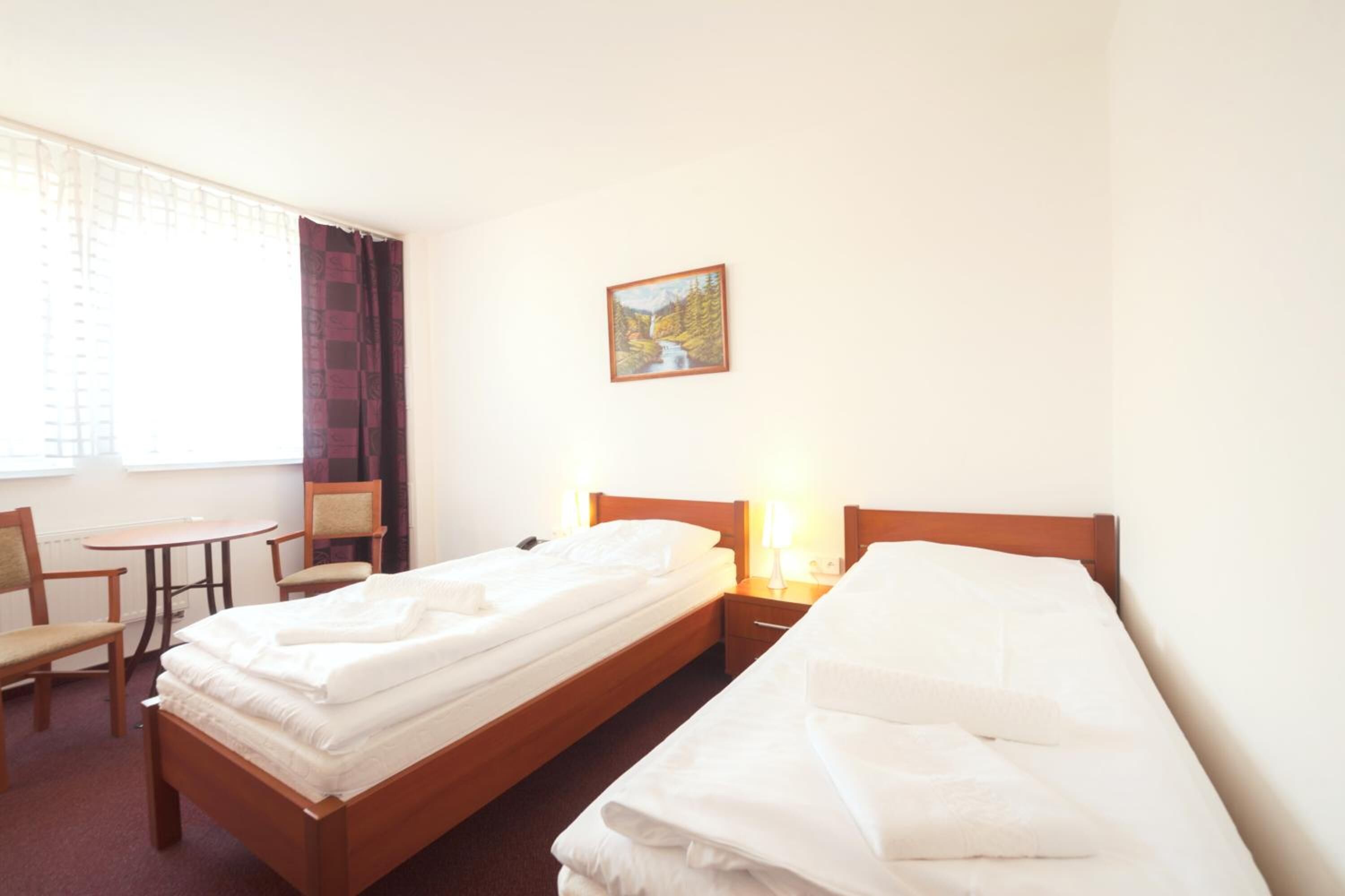 A hotel room with two single beds, a small table, a chair, and a painting on the wall.