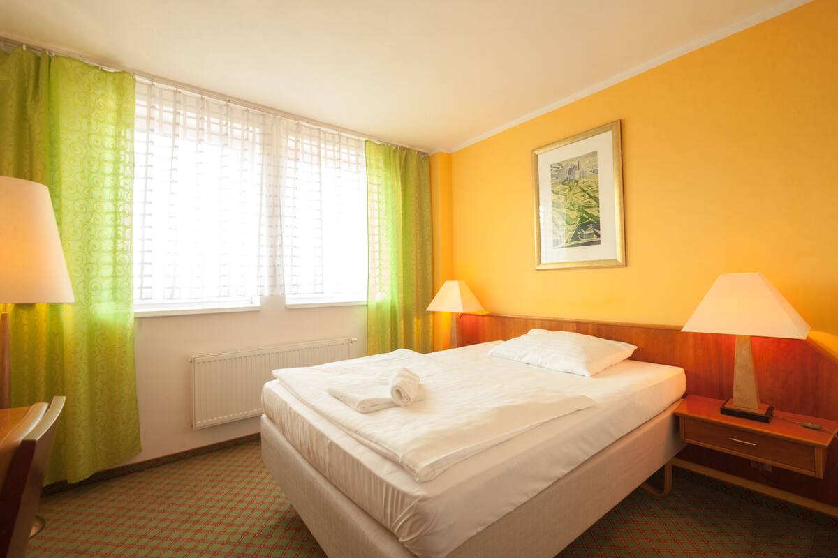Habitación de hotel con cama, dos lámparas de noche, ventana con cortinas verdes, radiador y un cuadro en la pared.