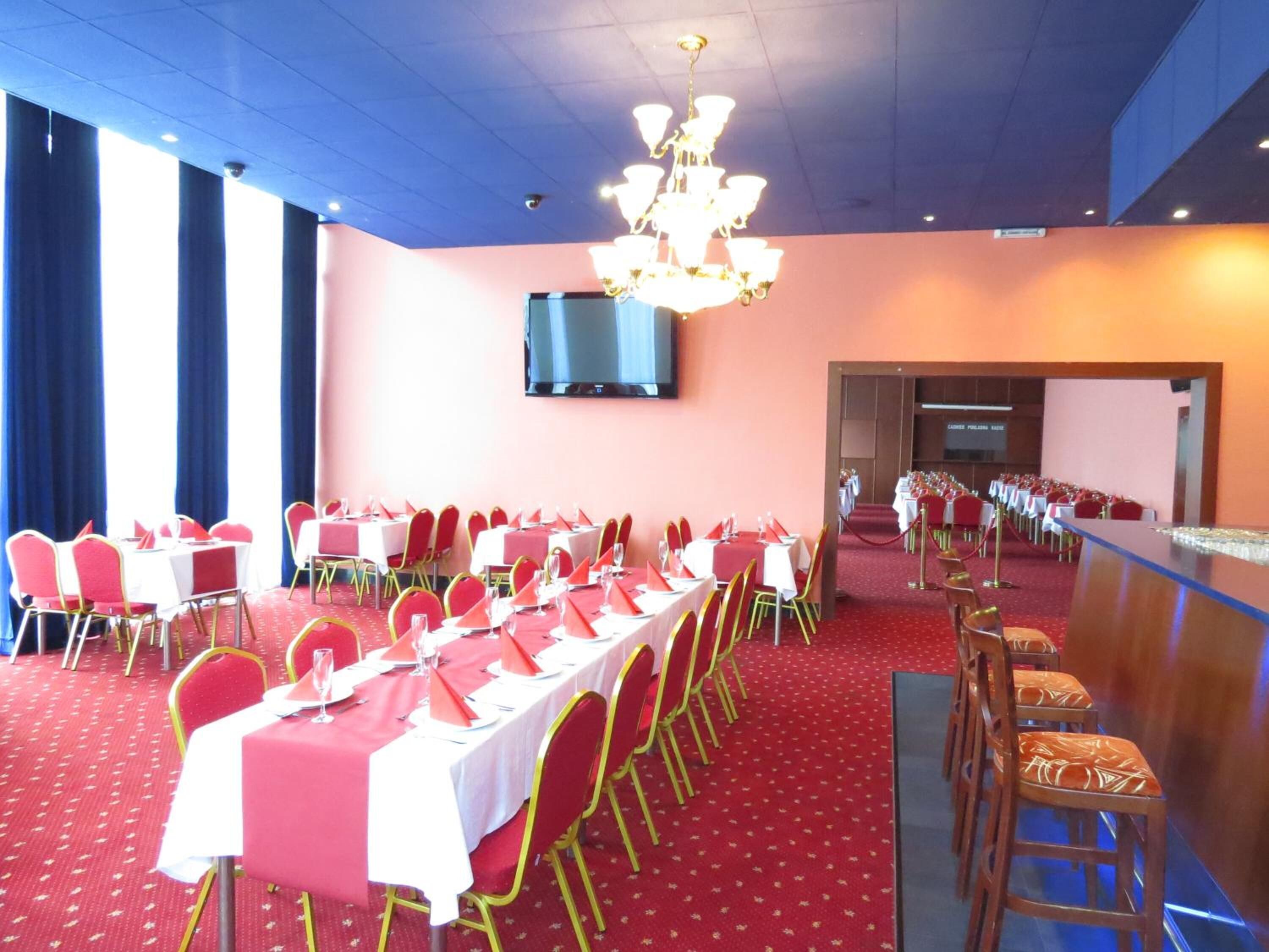 Une salle de banquet avec des tables dressées pour un événement formel, un lustre et un téléviseur à écran plat accroché au mur.