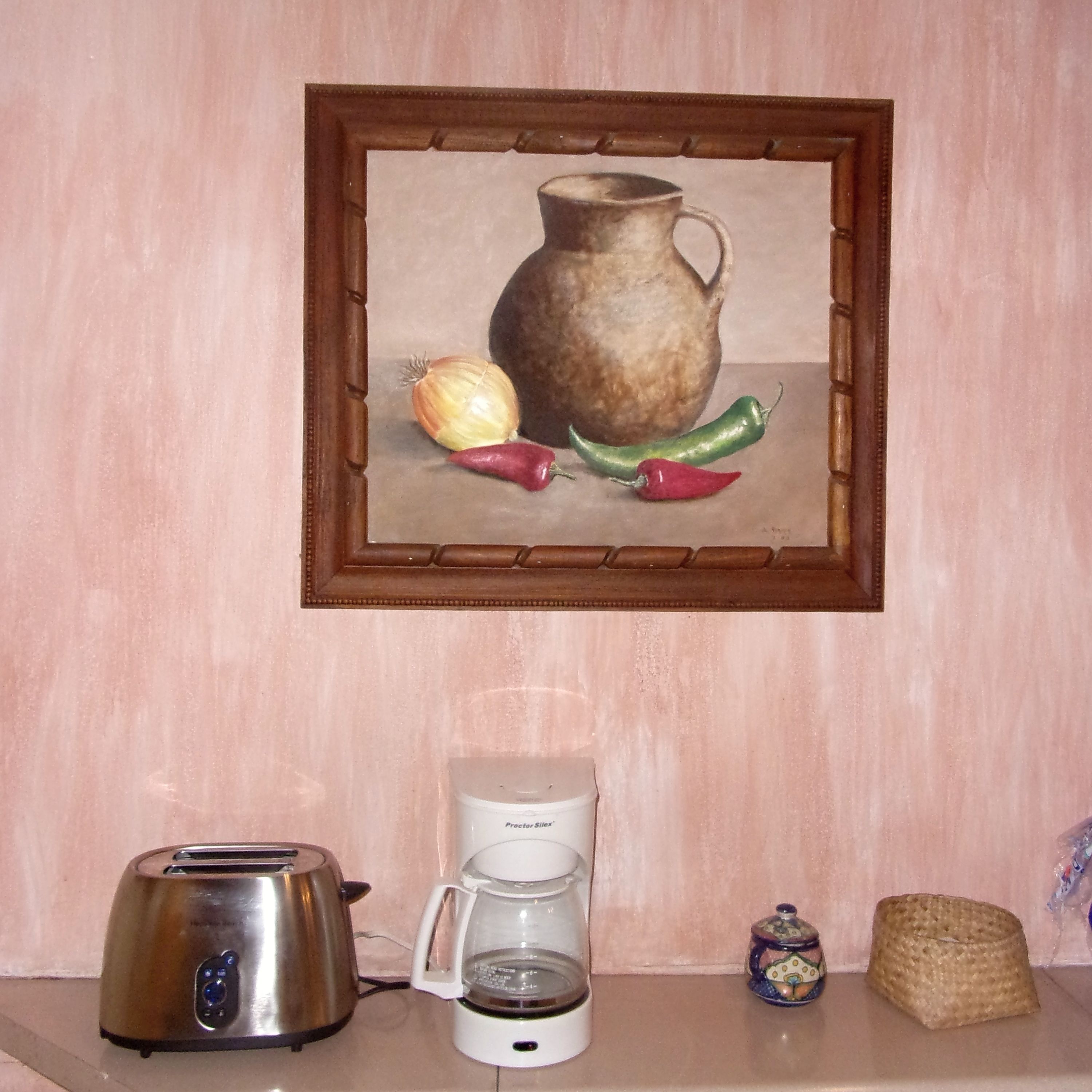 A kitchen counter with a toaster, a coffee maker, a framed painting of vegetables, and a small jar.