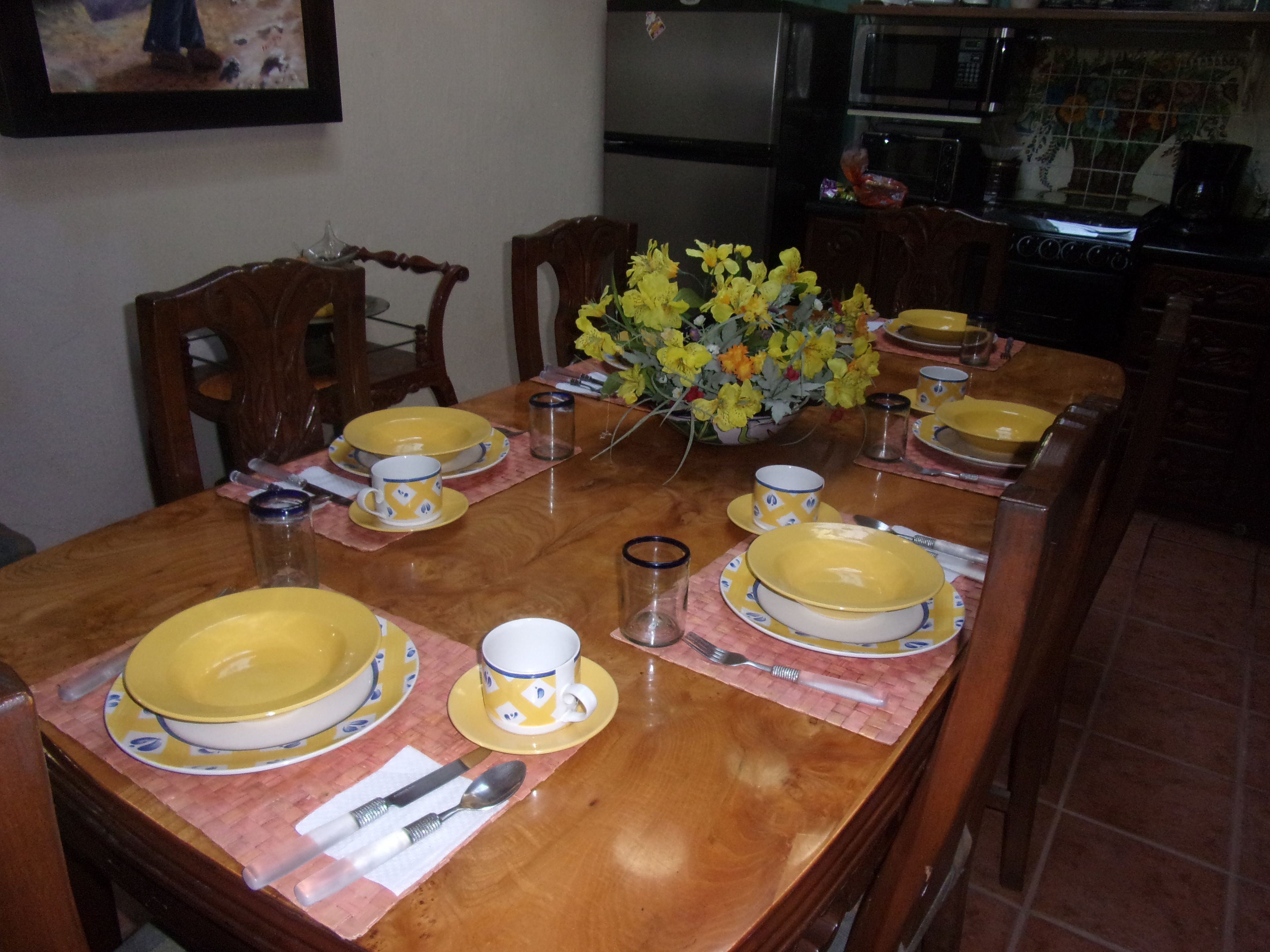 Una mesa de comedor dispuesta con platos, tazas y cubiertos amarillos, rodeada de sillas.