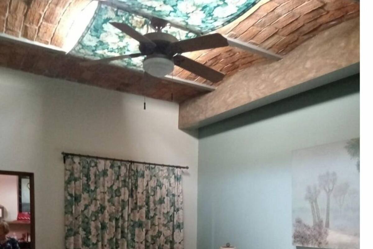Ein Schlafzimmer mit einem Bett, einem Deckenventilator, einem blumigen Vorhang, einem Stuhl und einem Bild an der Wand.