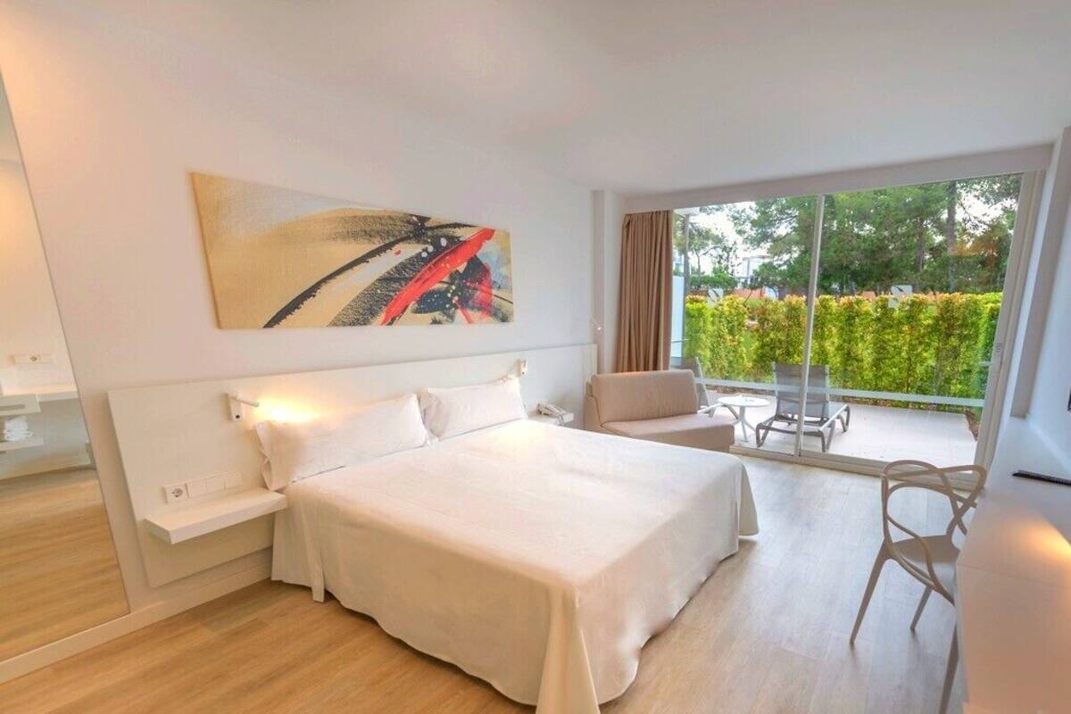 Una habitación de hotel moderna con una cama grande, un pequeño sofá, una mesa de comedor y un balcón con vistas a la vegetación.
