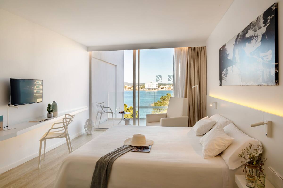 Una habitación de hotel moderna con una cama grande, televisión, balcón con vistas al mar y zona de comedor.