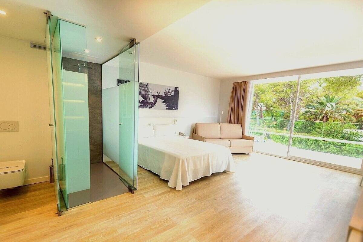 Un dormitorio moderno con cabina de ducha de vidrio, cama, sofá y un amplio ventanal con vistas a zonas verdes.
