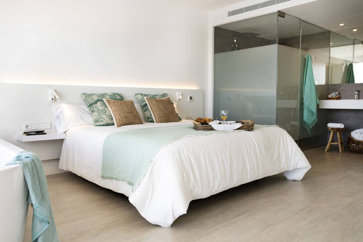 Un dormitorio moderno con una cama grande, mesitas de noche, un box de ducha de vidrio y un taburete pequeño.