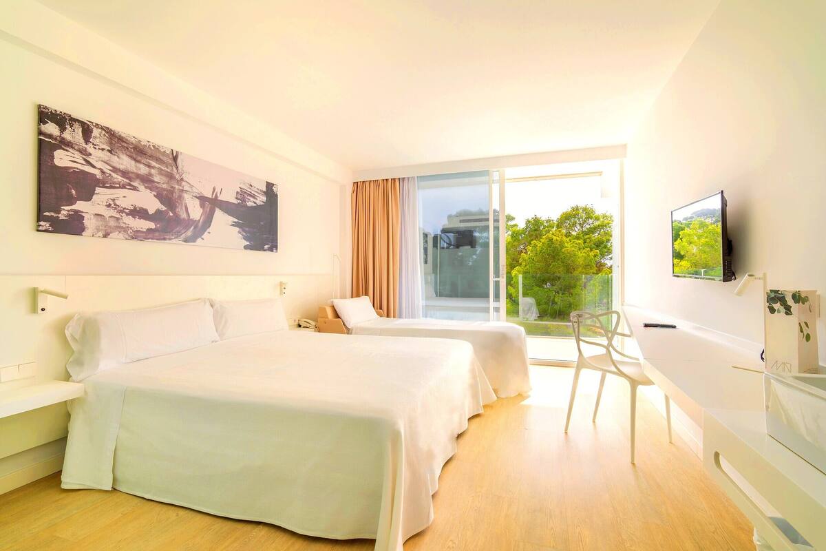 Habitación de hotel con dos camas, un escritorio, una silla, un televisor y un ventanal con vistas a árboles.