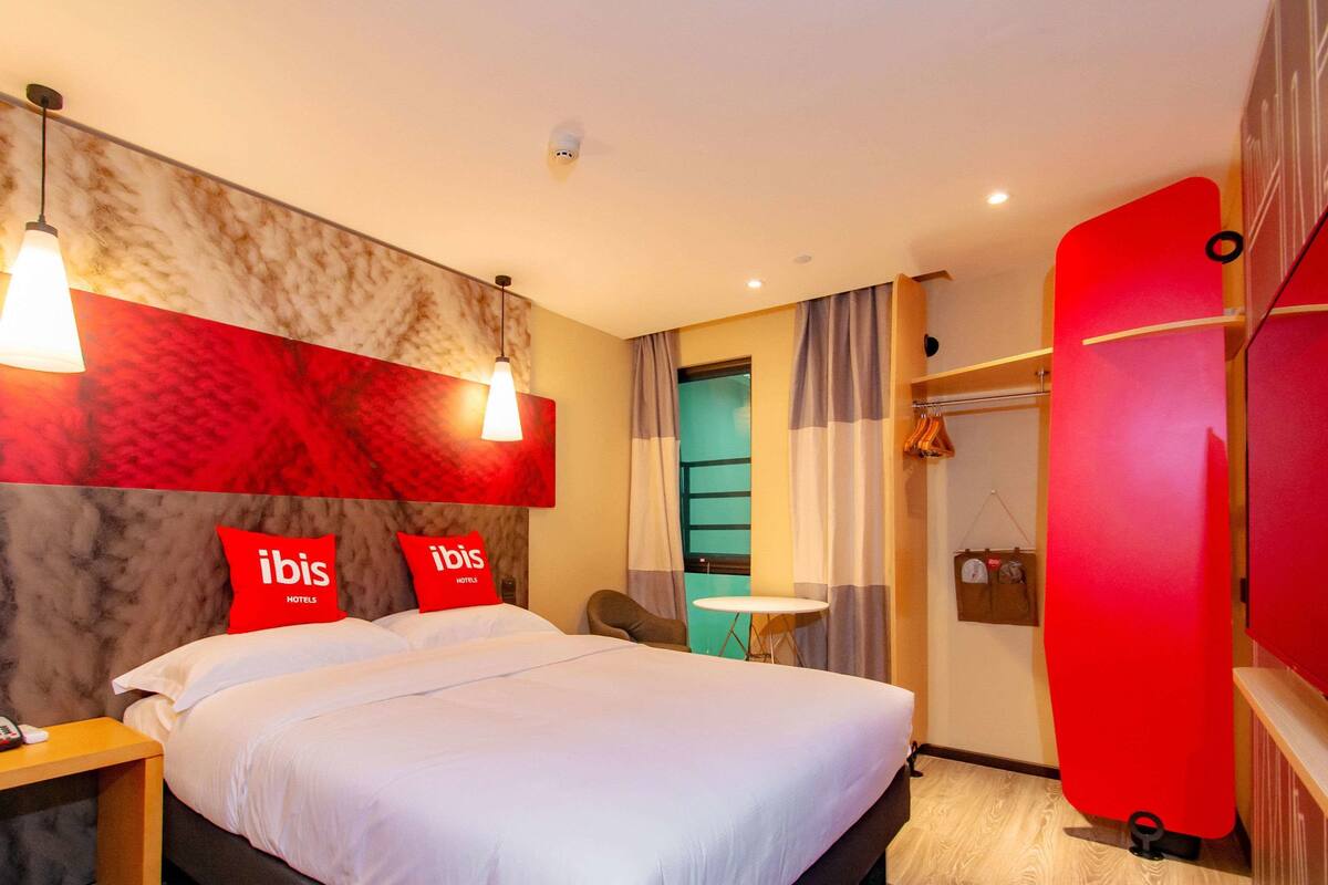 Une chambre d’hôtel avec un lit, deux oreillers rouges, une table de chevet, une table et un mur rouge.