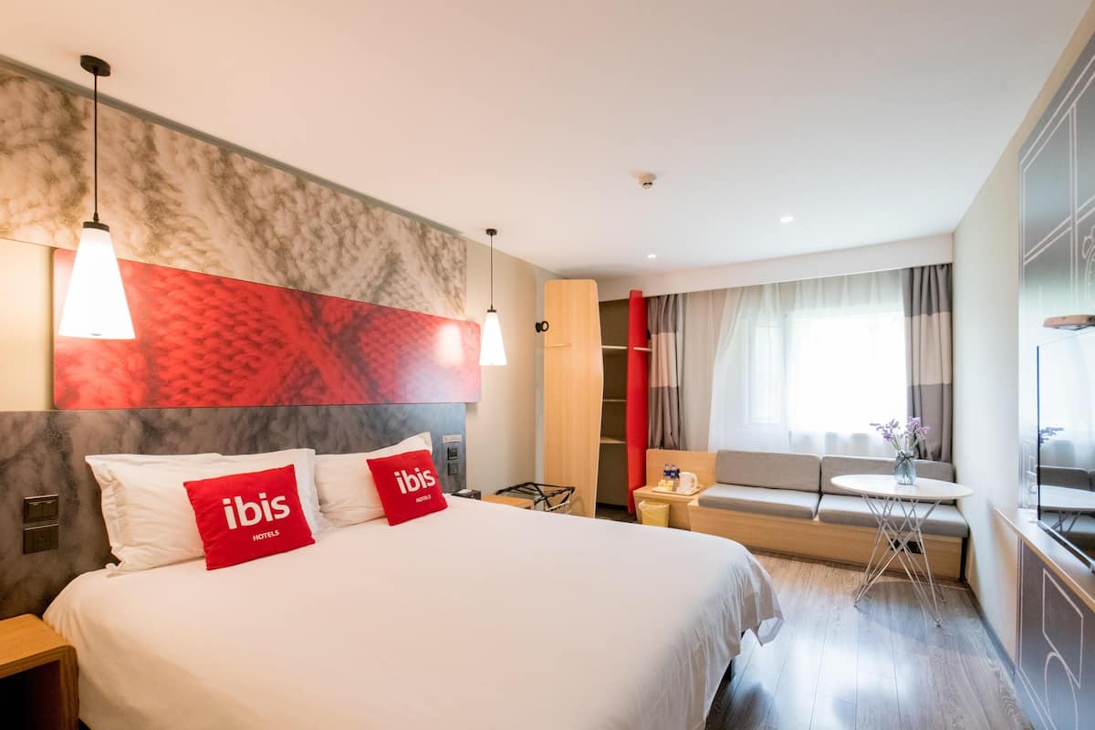 Une chambre d’hôtel avec un grand lit, deux tables de chevet, un bureau, une chaise, un canapé, une fenêtre avec des rideaux et un téléviseur fixé au mur.