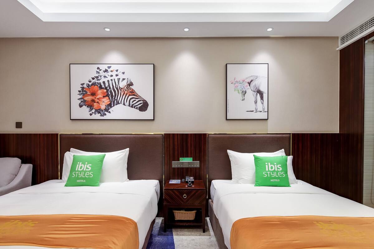 A hotel room with two beds, a nightstand, a wall-mounted control panel, and artwork above the beds.