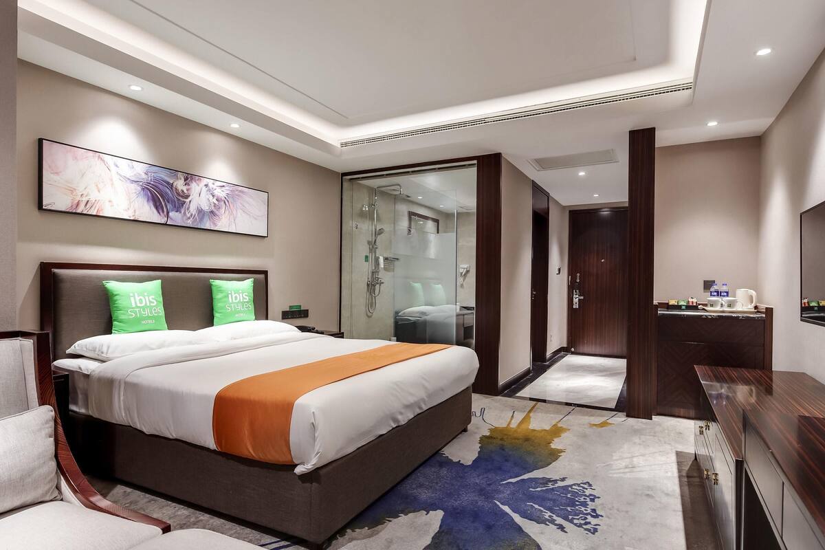 A modern hotel room with a large bed, a glass shower enclosure, a wooden nightstand, and a flat-screen TV.