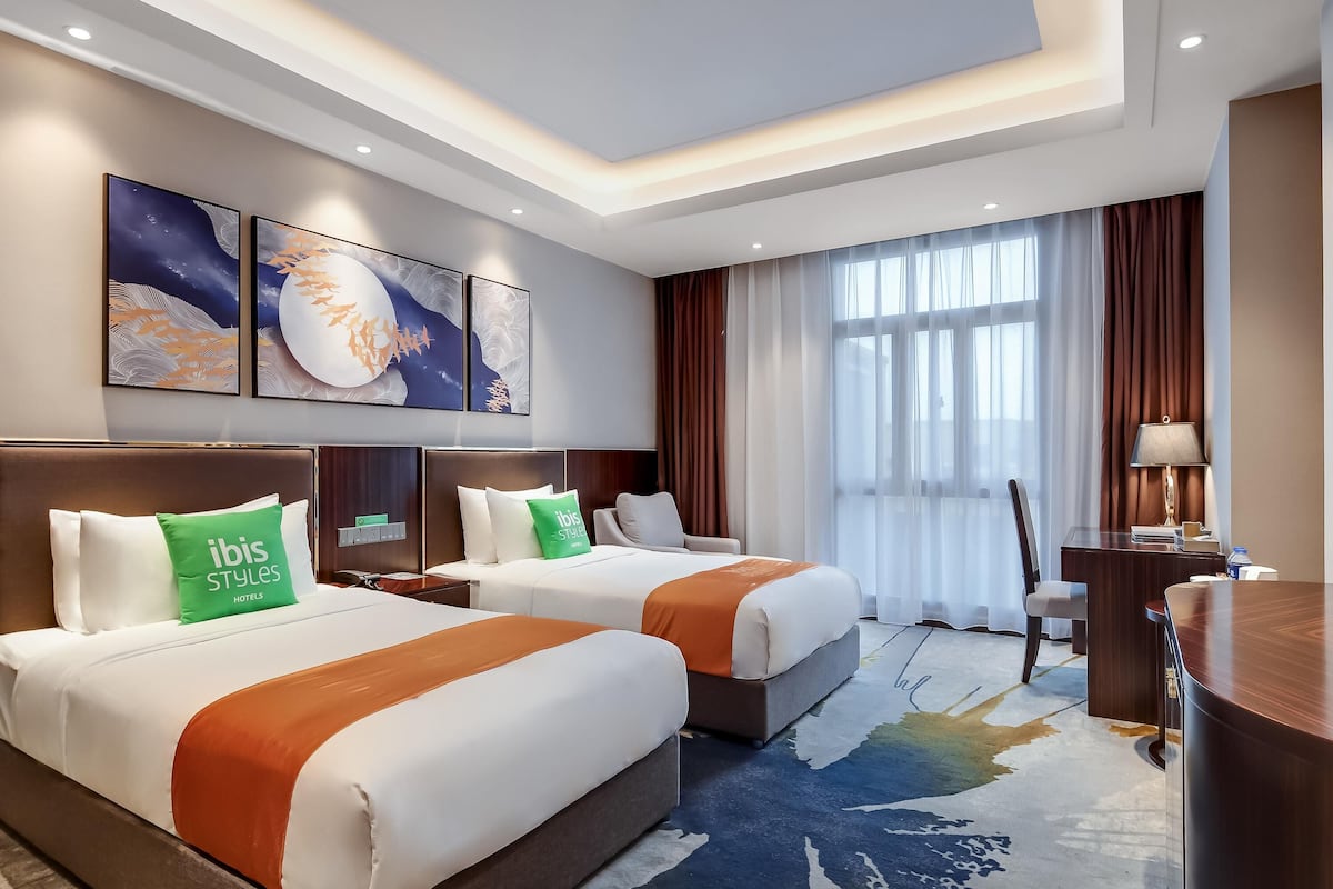 A hotel room with two beds, a desk, a chair, and a large window with curtains.