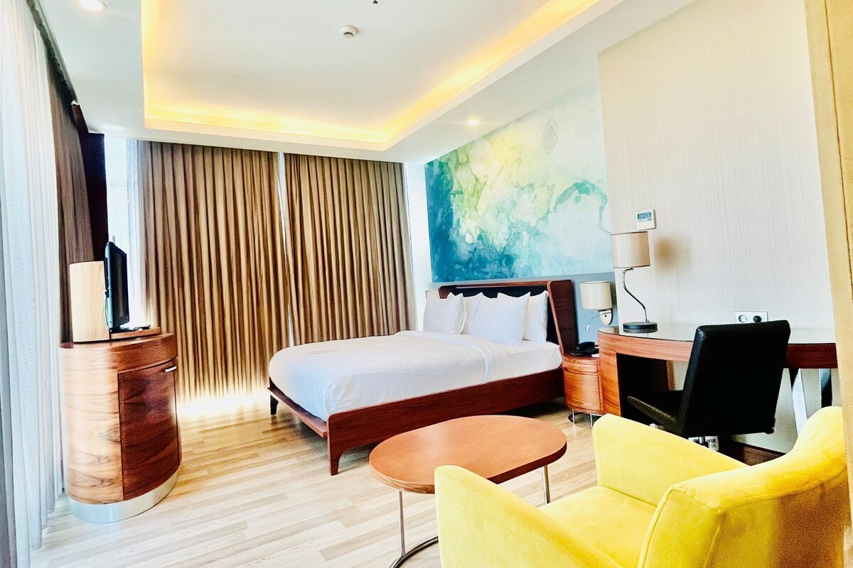 A hotel room with a bed, a desk, a chair, a lamp, and a painting on the wall.
