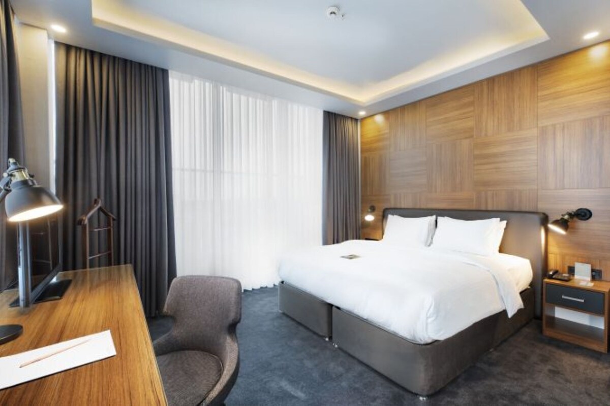 A modern hotel room with a large bed, a desk with a lamp, a chair, and a nightstand.
