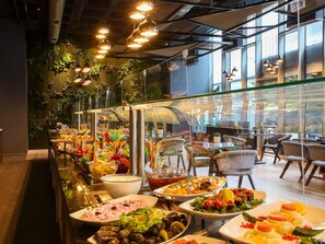 A buffet with various food items, a seating area with chairs, and a large window with a view of greenery. - Mercure Istanbul Umraniye (Istanbul)