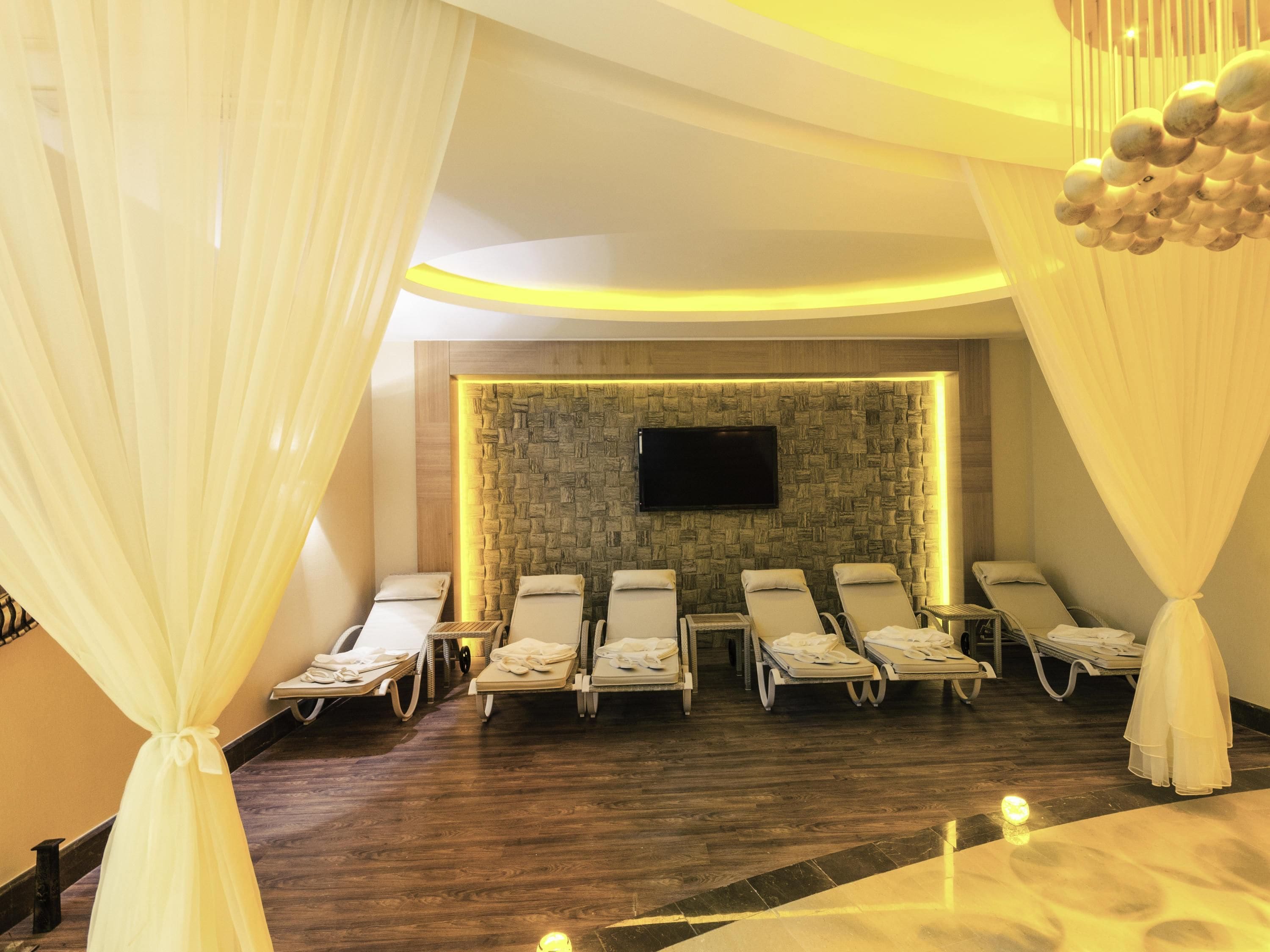 A spa room with wooden flooring, a stone wall, a flat-screen TV, and six sun loungers with towels.