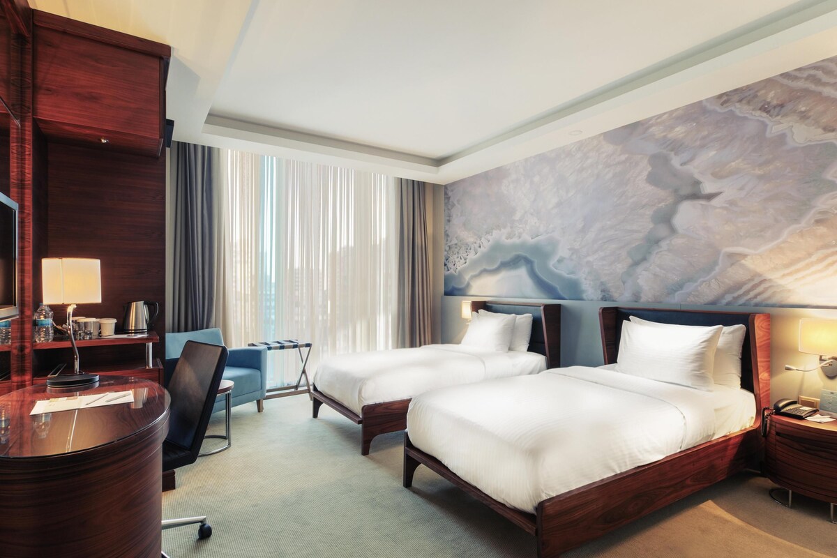 A hotel room with two beds, a desk, a chair, a large window with curtains, and a wall mural.