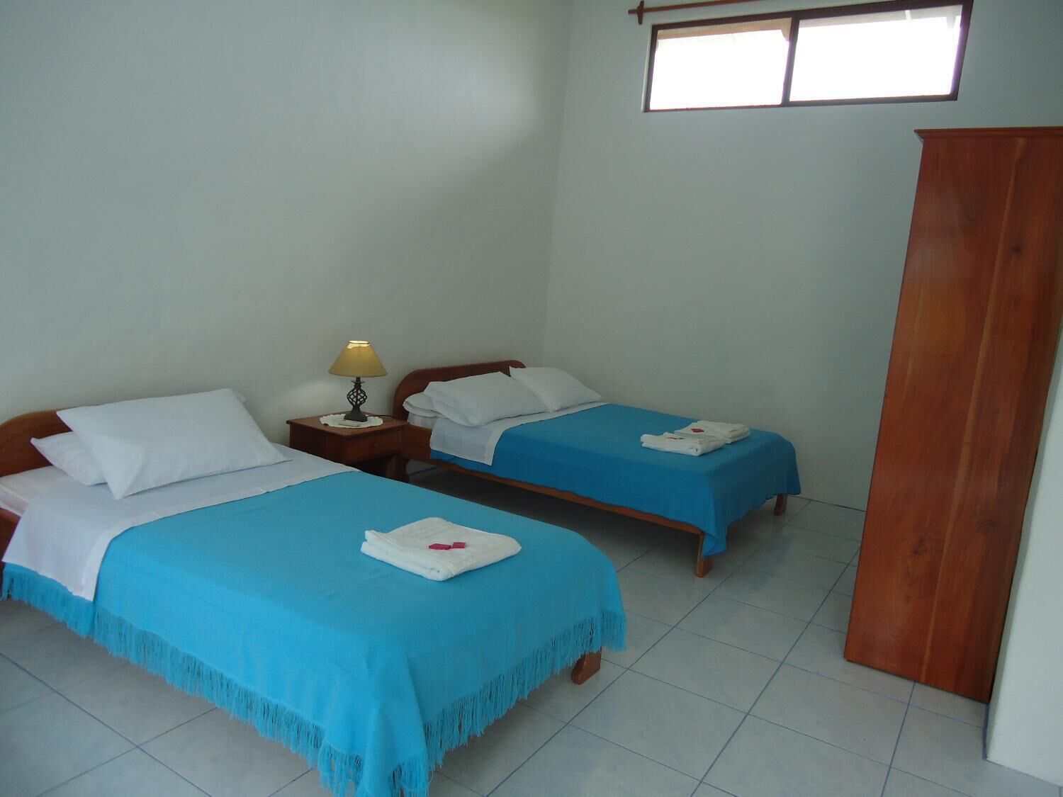 Una habitación con dos camas, cada una con una colcha azul y almohadas blancas. Hay una mesita de noche con una lámpara entre las camas.