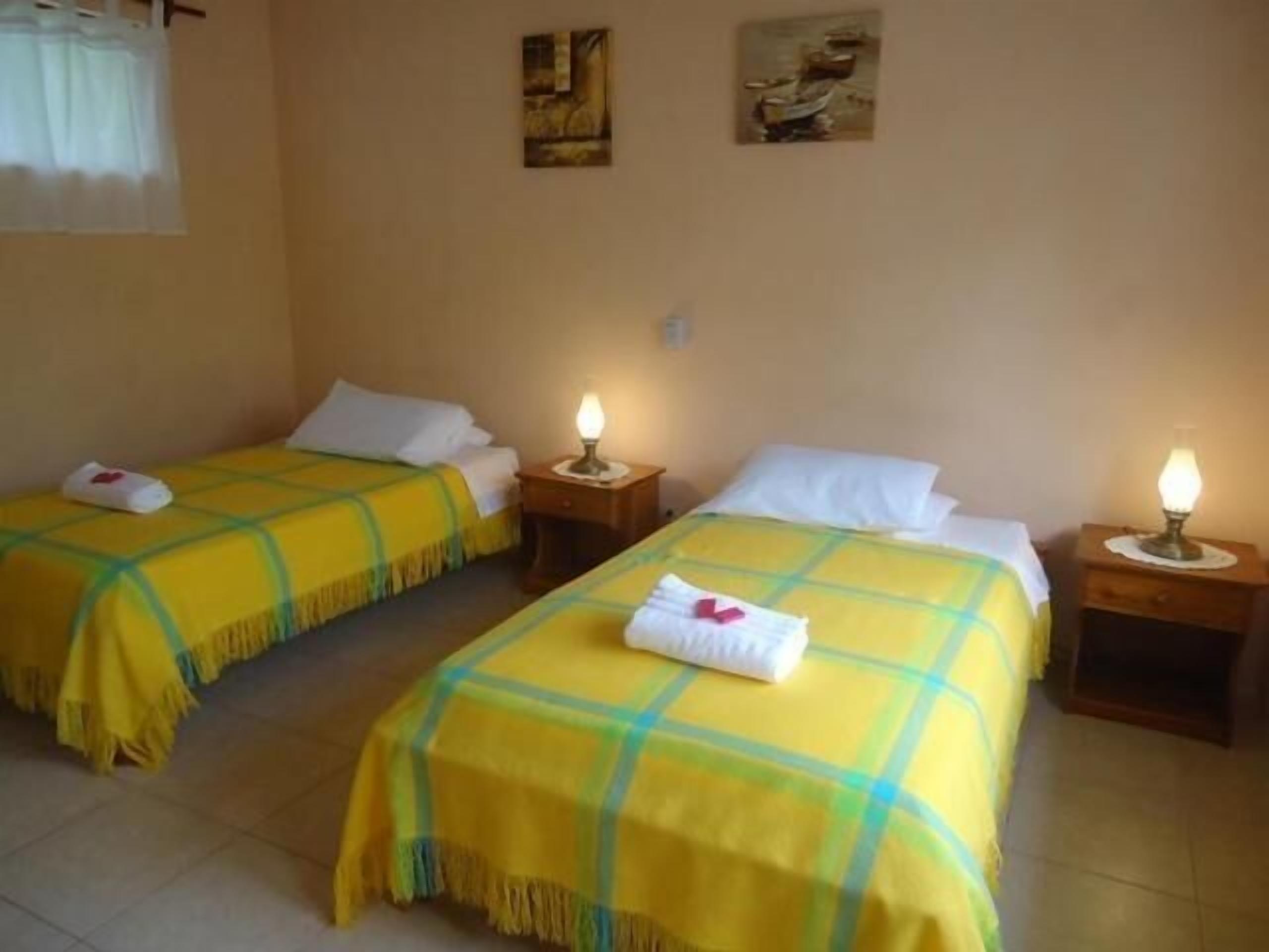 Habitación de hotel con dos camas individuales, cada una con colcha a cuadros amarillos y verdes, almohadas blancas y una mesita de noche con lámpara.