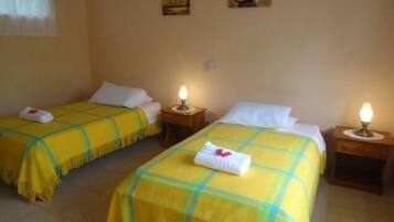 Habitación de hotel con dos camas individuales, cada una con colcha a cuadros amarillos y verdes, almohadas blancas y una mesita de noche con lámpara.