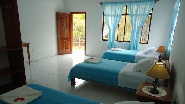 Una habitación con dos camas, una puerta de madera, un ventanal con cortinas azules y una mesita de noche con lámpara.