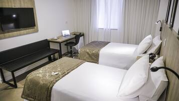 Ein Hotelzimmer mit zwei Betten, einem Schreibtisch mit einem Laptop, einem Fernseher und einem Fenster mit Vorhängen.