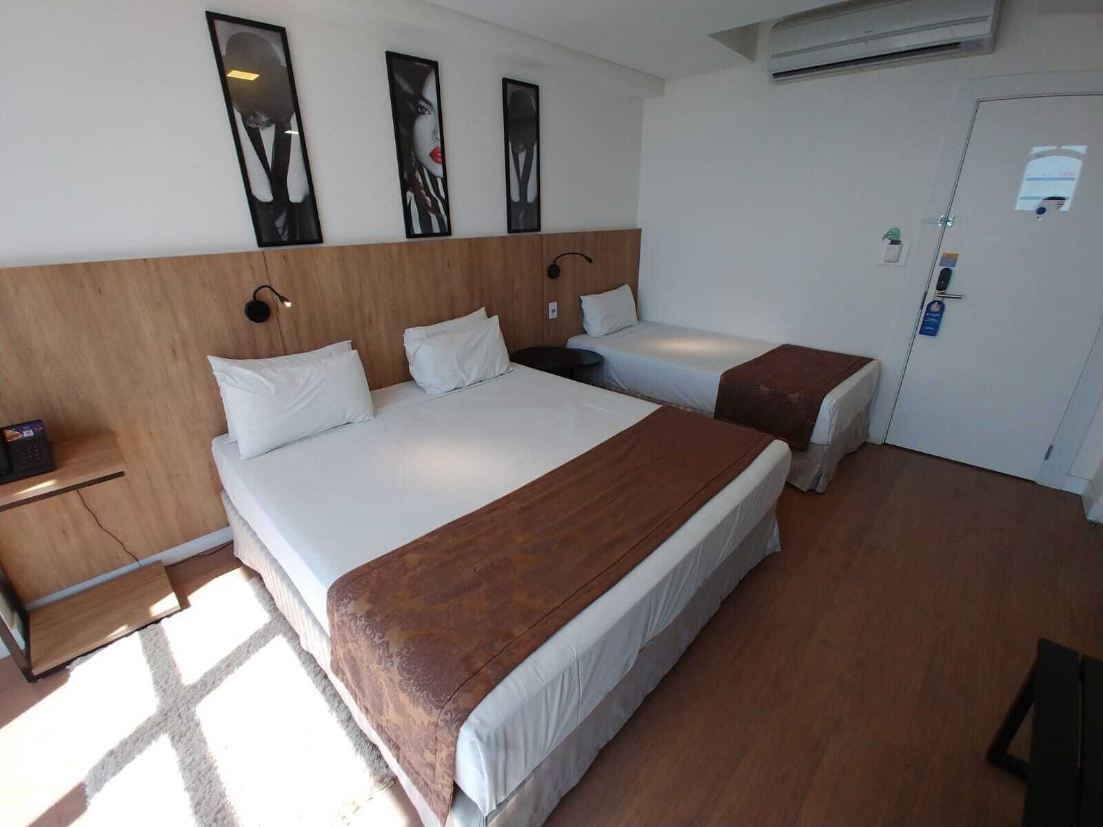 Habitación de hotel con dos camas, cabecera de madera, mesita de noche pequeña y aire acondicionado instalado en la pared.