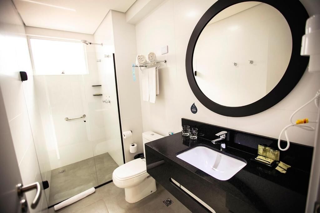 A modern bathroom with a glass-enclosed shower, a white toilet, a black sink with a white basin, and a large round mirror.