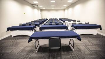 Uma sala de conferências com mesas e cadeiras dispostas em fileiras, piso coberto de carpete e paredes brancas.