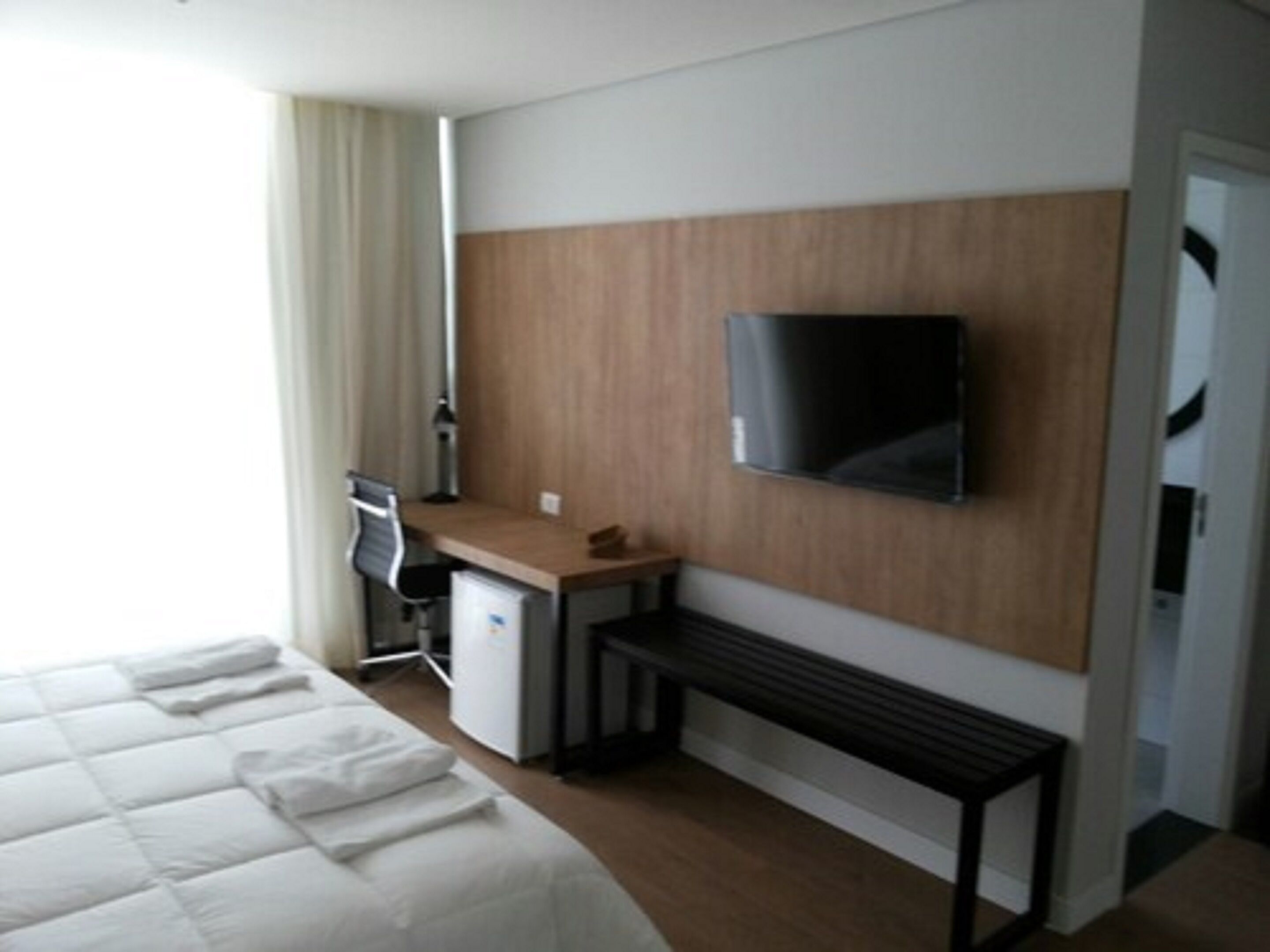 A hotel room with a bed, a desk, a chair, a television mounted on the wall, and a small table.