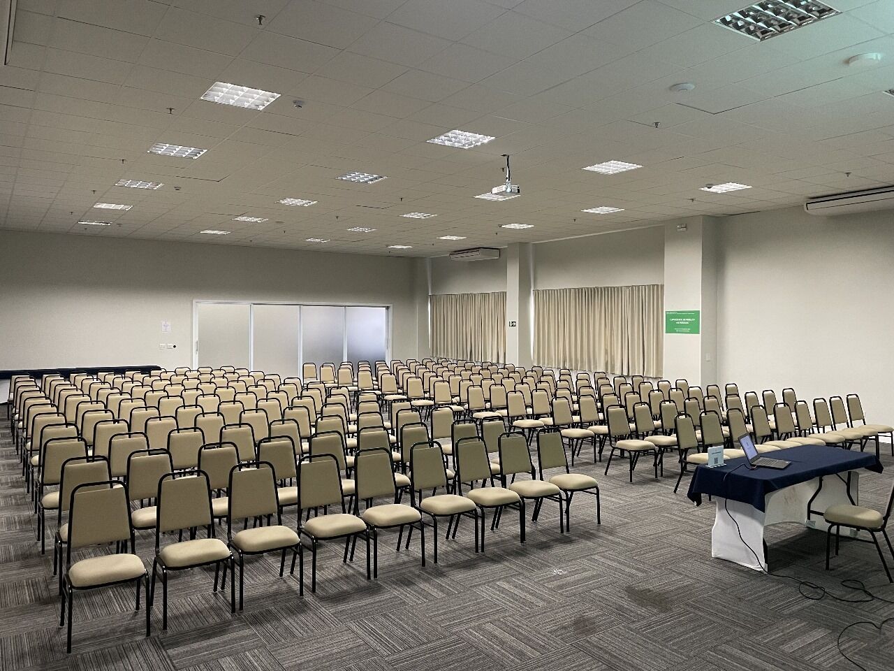 Un salón de conferencias con filas de sillas, una pantalla de proyector y una mesa con una computadora portátil.