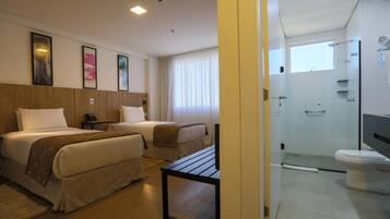 Quarto de hotel com duas camas, uma mesinha, televisão, chuveiro e sanitário.