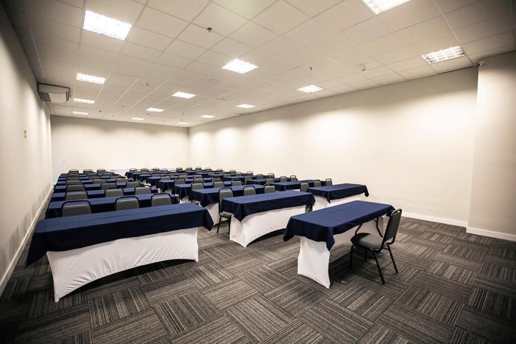 Una sala conferenze con file di tavoli e sedie, pavimento in moquette e soffitto bianco con luci fluorescenti.