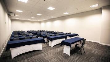 Una sala conferenze con file di tavoli e sedie, pavimento in moquette e soffitto bianco con luci fluorescenti.