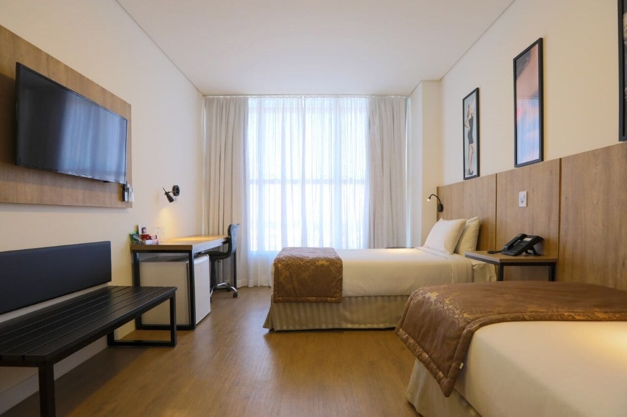 Quarto de hotel com uma cama, uma escrivaninha, uma cadeira, televisão na parede e uma janela com cortinas.