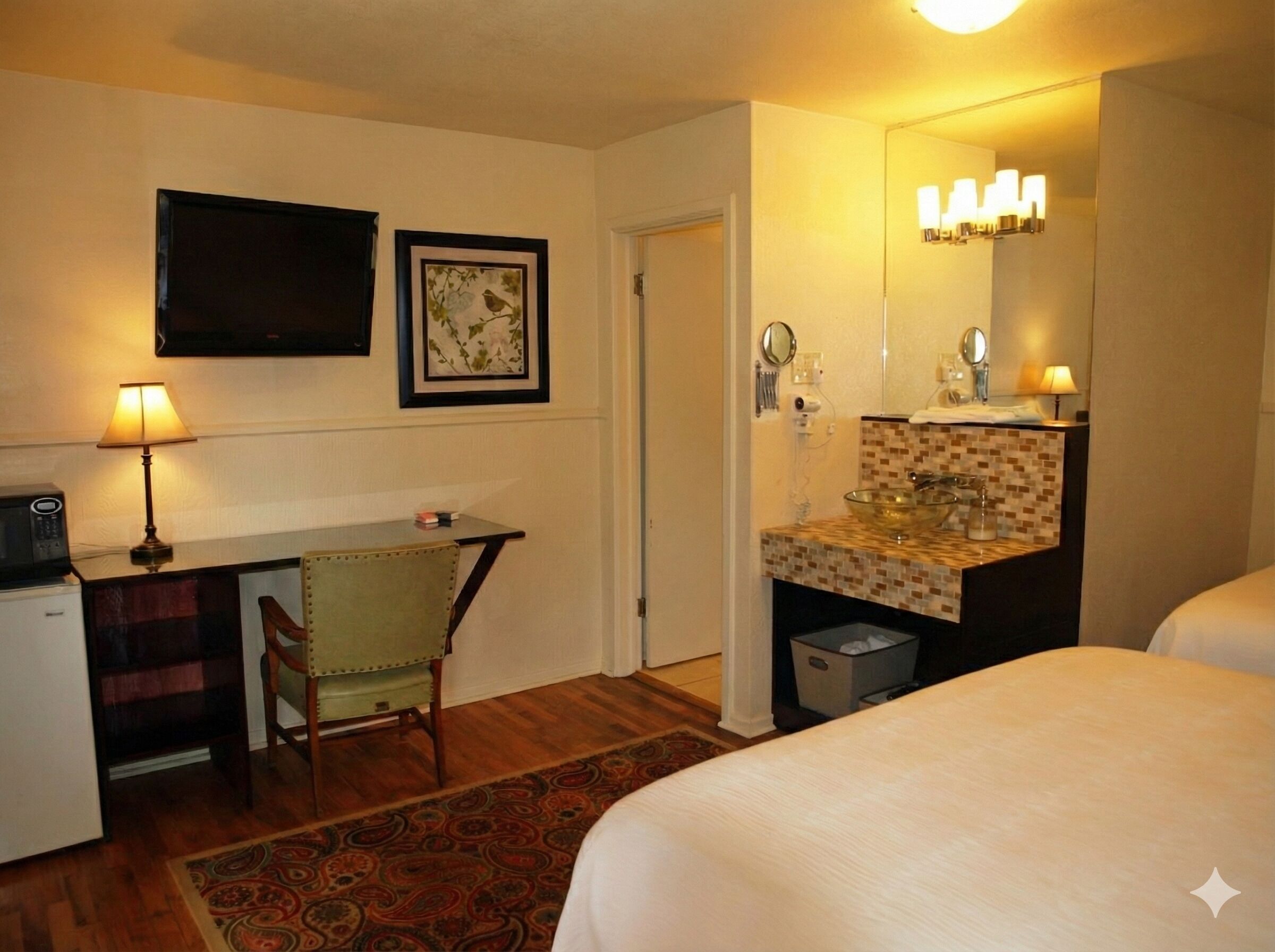 A hotel room with a bed, a desk with a lamp, a chair, a bathroom with a sink and mirror, and a television on the wall.