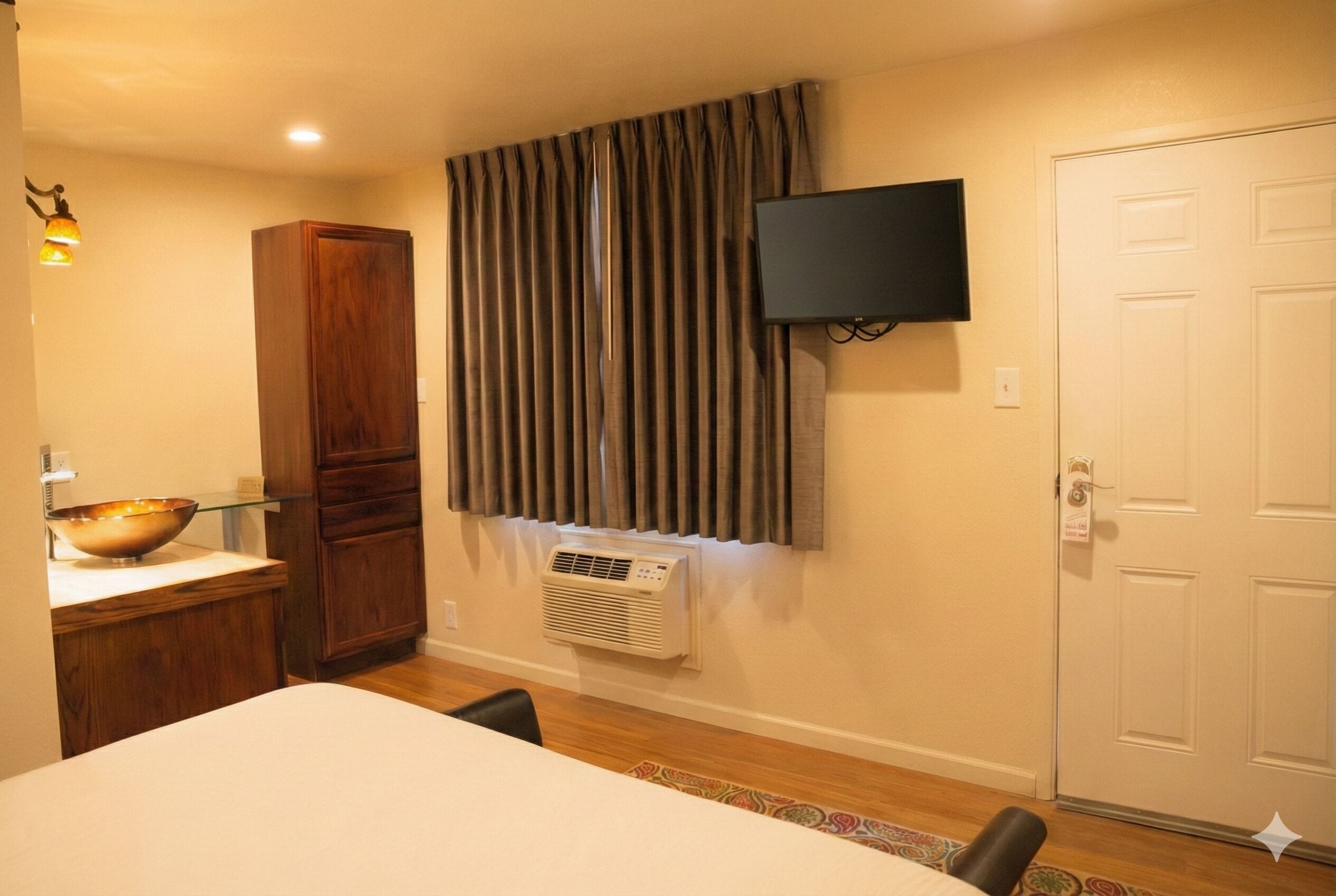 A hotel room with a bed, a wooden cabinet, a television mounted on the wall, and a window with curtains.