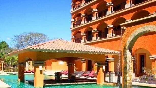 Un hotel con zona de piscina, que cuenta con un edificio de varios pisos con balcones y un área de descanso techada.