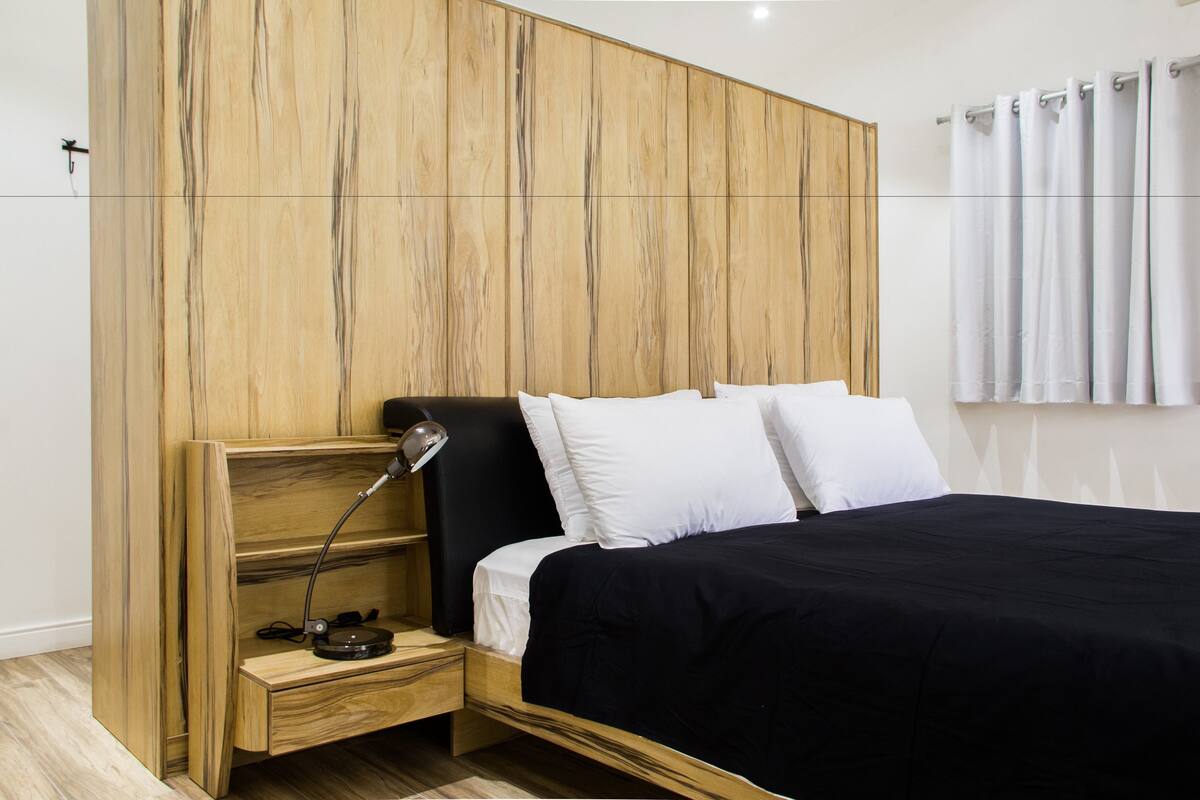 A modern bedroom with a wooden headboard, a black bedspread, white pillows, and a bedside lamp.