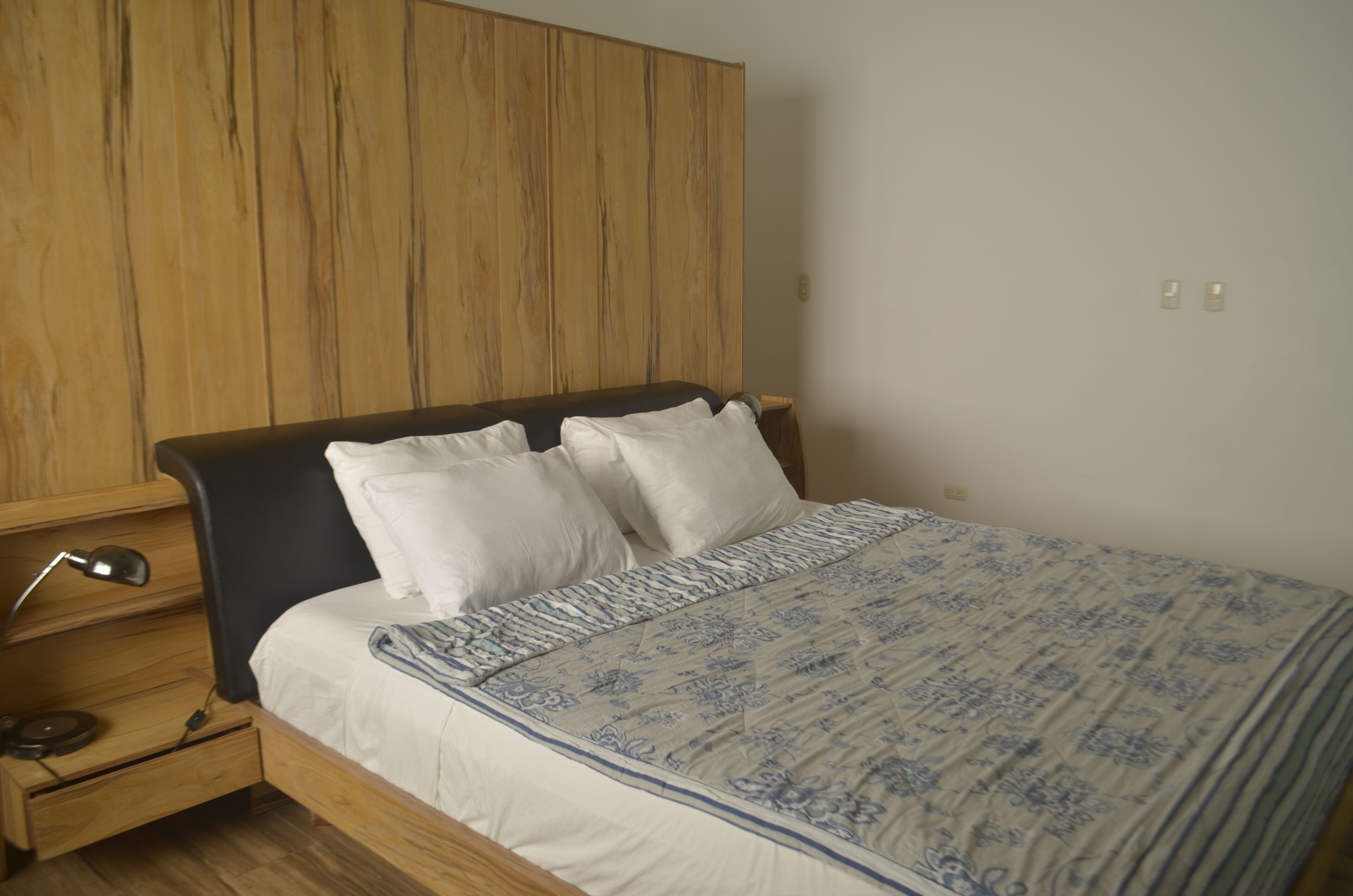 Quarto com cabeceira de madeira, cama com roupa de cama branca e azul, mesa de cabeceira com abajur e uma gaveta.