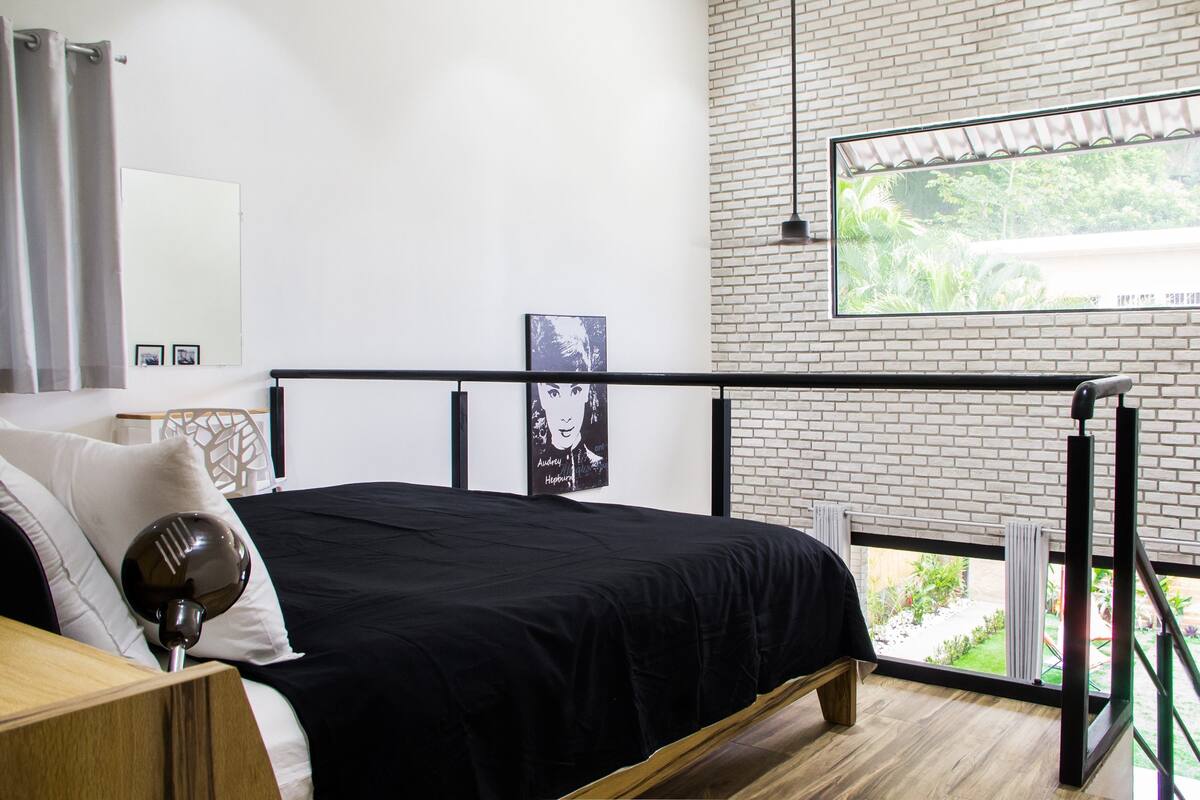 A bedroom with a black bed, a wooden nightstand, a white wall, a black railing, and a window with a view of greenery.
