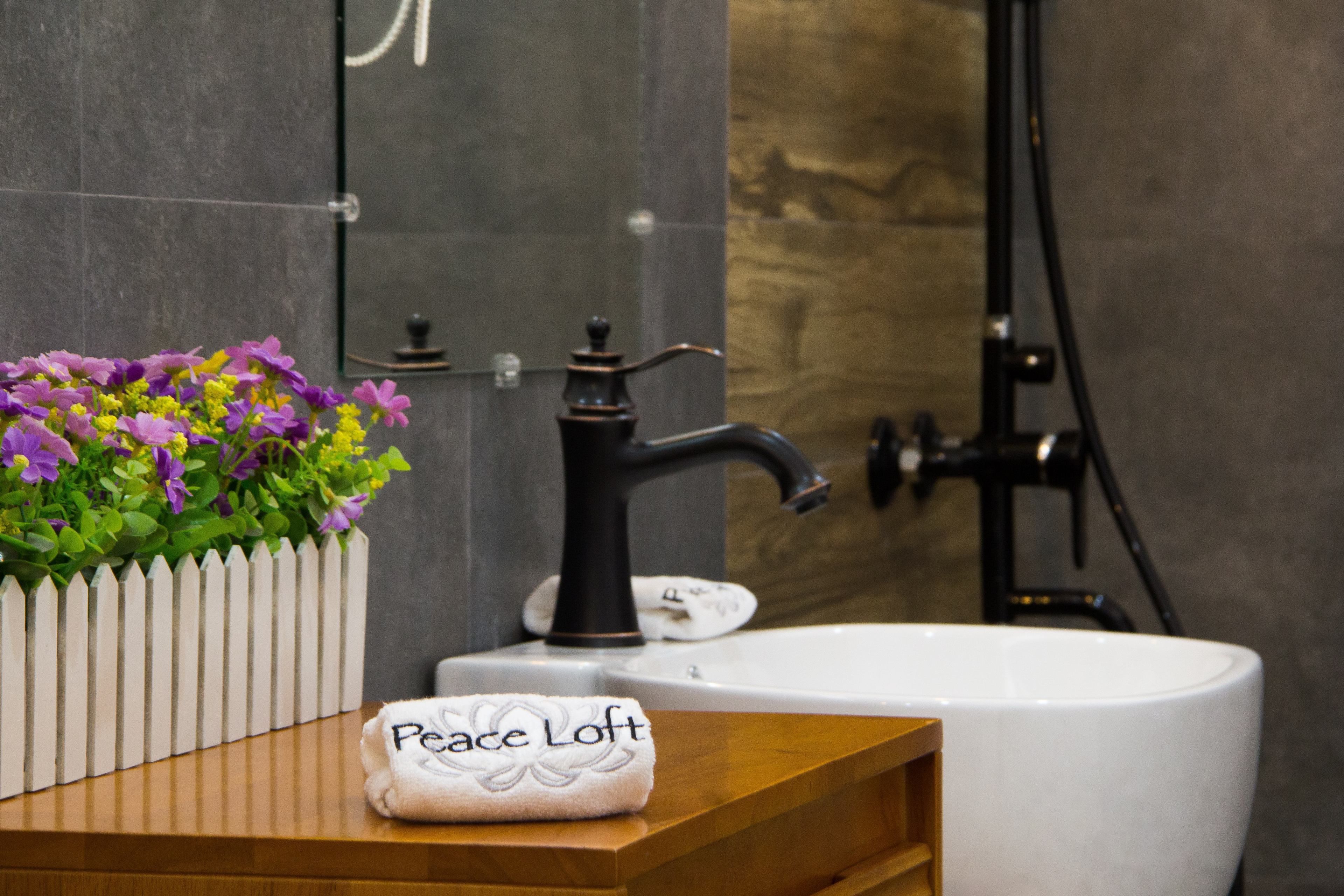 A bathroom with a white sink, a black faucet, a white towel with "Peace Loft" embroidered on it, and a planter with purple and green flowers.