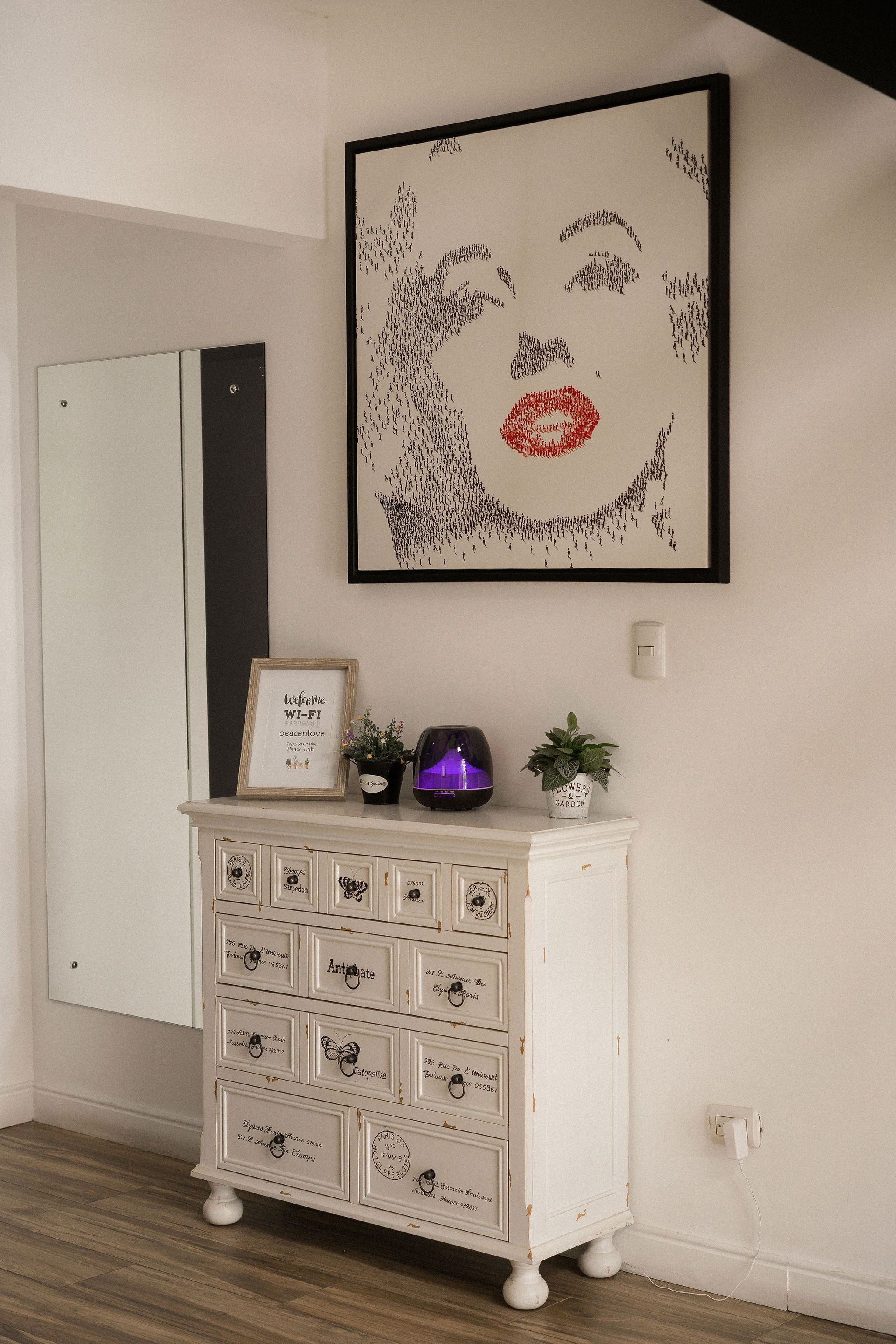A white dresser with multiple drawers, a framed picture with a red lipstick design, and a small potted plant on top.