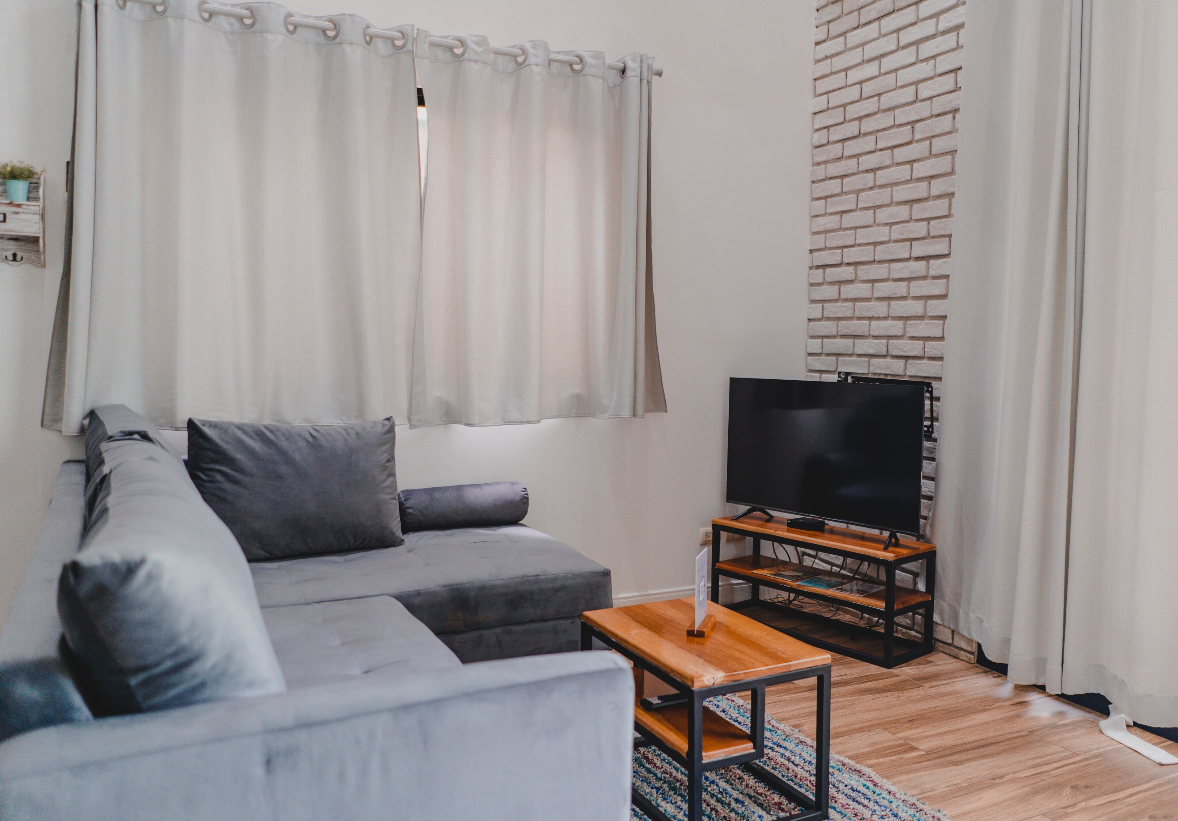 A living room with a grey sofa, a wooden coffee table, a flat-screen TV on a wooden stand, and a brick wall.