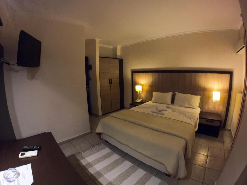 A hotel room with a bed, bedside lamps, a flat-screen TV mounted on the wall, a desk with a remote control, and a check-in form.