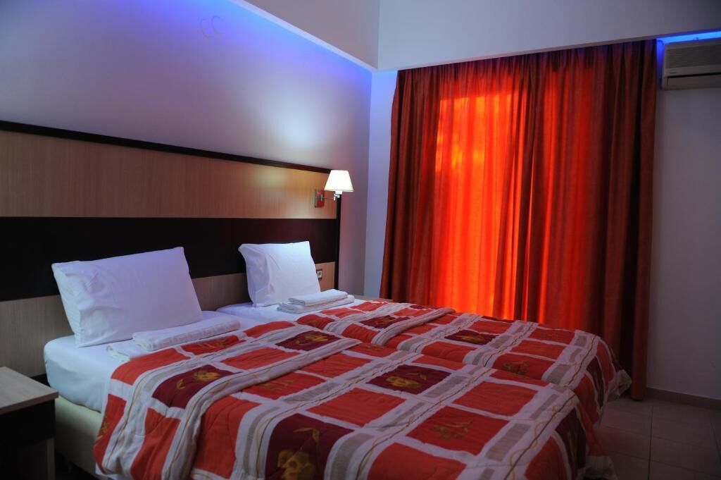 A hotel room with a bed, a nightstand, a lamp, and a window with red curtains.