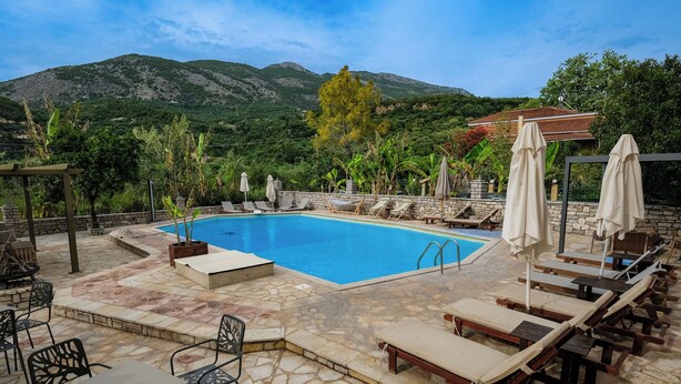 Zona de piscina con sillones de descanso y sombrillas, rodeada de muros de piedra y vegetación, con montañas al fondo.