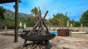 Un pozo de fuego con leña, rodeado de baldosas de piedra, con una piscina y sillones de descanso al fondo.