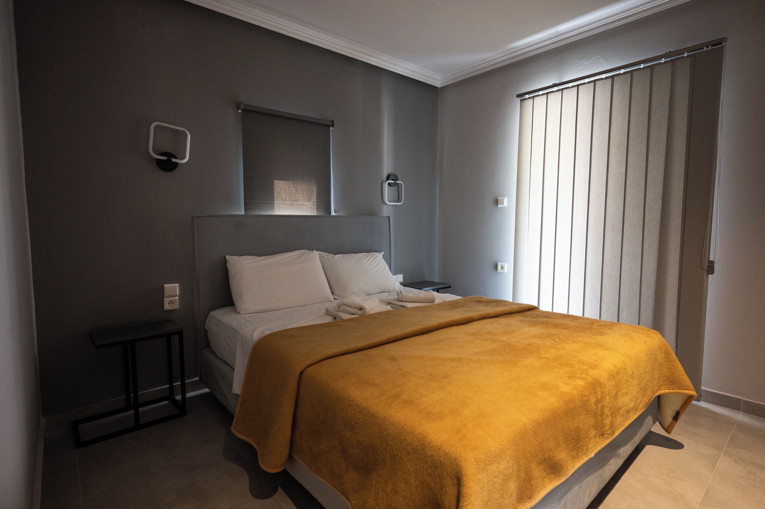 Un dormitorio con cama, mesita de noche, lámpara de pared y ventana con persianas.
